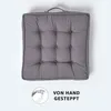 A square gray cushion with tufted buttons and a handle on top, set against a white background. A small circular graphic with a hand sewing and the text "VON HAND GESTEPPT" is in the bottom left corner. Náhled obrázku produktu