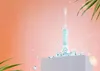 A blue electric toothbrush with a lion design on a white pedestal, surrounded by water splashes and green leaves, against a peach background. Produktbild-Vorschau