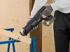 A person wearing gloves is using a cordless reciprocating saw to cut a wooden beam supported by sawhorses. Produktbild-Vorschau