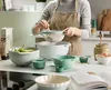 A person is sifting flour into a mixing bowl in a kitchen setting, surrounded by various kitchen utensils. Produktbild-Vorschau