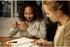 Two young women are sitting at a table, one holding a cup of tea and looking at a notebook, while the other points at something on the table. Anteprima dell’immagine prodotto