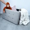 A person's hand is placing a stuffed orangutan into a gray storage ottoman. A pink pillow and a white fluffy blanket are also inside. Produktbild-Vorschau
