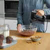 A person is using a black Bosch hand mixer to mix batter in a glass bowl, with muffins and ingredients on a wooden table. Produktbild-Vorschau