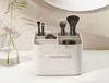 A makeup organizer with brushes and a compact is shown on a white surface with a plant in a vase to the left. Náhled obrázku produktu