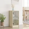 A small cabinet with glass doors is shown in a room with a plant and a painting on the wall. Náhľad obrázku produktu