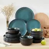 A collection of teal and black stoneware dinnerware is arranged on a light-colored wooden surface, with a vase of dried flowers and a decorative wooden arch in the background. Náhled obrázku produktu