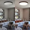 A split image showing a living room with a sectional sofa, coffee table, and decorative mirrors. The left side shows the room with the ceiling light off, and the right side shows the room with the ceiling light on. Produktbild-Vorschau