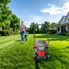 A woman is pushing a wheelbarrow on a lawn next to a lawnmower, with houses in the background. Náhled obrázku produktu