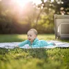 A baby is crawling on a blanket on the grass in a park with a travel crib in the background. Podgląd zdjęcia produktu