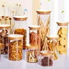 A collection of glass storage jars with bamboo lids, filled with various dry goods like pasta, nuts, crackers, and spices, arranged on a white surface. Produktbild-Vorschau