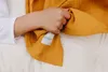 A child's hand holding a mustard yellow muslin blanket with a brand tag. Produktbild-Vorschau