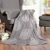 A gray blanket with a circular pattern is draped over a gray armchair in a living room setting. Books are stacked on the floor next to the chair, and a small ottoman with a tray of snacks is visible in the background. Produktbild-Vorschau