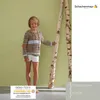 A young girl wearing a knitted sweater and shorts stands next to two birch trees against a light green wall. Produktbild-Vorschau