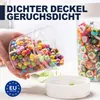 A hand is holding a clear plastic container filled with colorful candies, tilted to show the contents. Another clear container filled with colorful candies is visible in the background, along with a white lid. A blue EU logo is in the bottom left corner. Produktbild-Vorschau