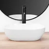 A white oval basin sink with a black faucet is shown in a studio setting with a black circular mirror in the background and a wooden surface in the foreground. Náhľad obrázku produktu