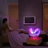 A woman is lying on a bed, wrapped in a blanket, next to a heart-shaped lamp that is lit up with colorful lights. Produktbild-Vorschau