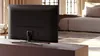 The back of a television is shown on a stand, with a living room in the background. Produktbild-Vorschau