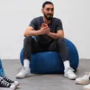 A man sits on a blue beanbag chair in a room with a white wall and gray floor. Several pairs of sneakers are visible around the beanbag. Voir les images du produit