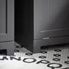 Close-up view of the bottom of two gray furniture pieces, possibly cabinets or a wardrobe, standing on a black and white hexagonal tiled floor. A portion of a white rug with black letters is visible in the foreground. Anteprima dell’immagine prodotto