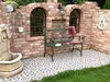 A garden bench is placed on a patterned patio in front of a brick wall with arched windows. A fountain is to the left, and grass is in the foreground. Anteprima dell’immagine prodotto