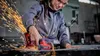 A woman is using a red and black cordless angle grinder to cut a piece of metal, with sparks flying. Produktbild-Vorschau