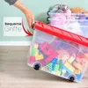 A hand is shown closing the lid of a transparent storage box filled with toys. Other similar boxes are stacked behind it. Produktbild-Vorschau