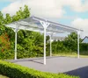 A white aluminum carport with a clear roof is set up on a paved driveway, surrounded by lush green bushes and trees under a partly cloudy sky. Podgląd zdjęcia produktu