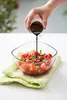 A hand is pouring dark liquid from a small metal container into a glass bowl filled with chopped tomatoes and herbs. Podgląd zdjęcia produktu