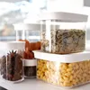 Several clear plastic food storage containers with white lids are arranged on a surface. The containers hold various dry goods like rice, pasta, star anise, and dried fruit. Náhľad obrázku produktu