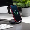 A red iPhone, Apple Watch band, and AirPods are charging on a multi-device wireless charging station on a desk next to a computer monitor and keyboard. Produktbild-Vorschau