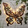A decorative metal butterfly with LED lights is mounted on a brick wall, surrounded by greenery. Produktbild-Vorschau