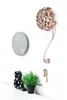 A display of decorative items on a white wall, including two clocks, an ampersand light, and a potted plant. Produktbild-Vorschau