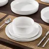 A table setting with white ceramic plates and bowls on a brown wooden table with a woven placemat. Produktbild-Vorschau