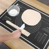 A person is using a black silicone baking mat with measurement markings to cut dough with a white knife. A rolling pin and a bowl of flour are also on the mat. Voir les images du produit