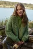 A young woman sits outdoors by a lake, wearing a green cardigan and shirt. Produktbild-Vorschau