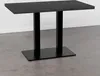 A dark marble-top bistrot table with a black metal pedestal base, shown in a studio setting with a light gray wall and a concrete floor. Produktbild-Vorschau