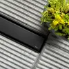 A close-up view of a black shower drain on a textured gray surface, with a potted plant in the upper right corner. Produktbild-Vorschau