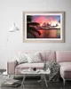 A framed picture of a sunset over the ocean is displayed on a wall above a pink sofa in a living room. Produktbild-Vorschau