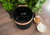 A modern rice cooker with a digital display sits on a wooden table, surrounded by plants and bowls of rice. Voir les images du produit