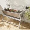A stainless steel electric rotisserie grill with vegetables on the grate, placed on a wooden deck next to a white wall with shadows. Produktbild-Vorschau