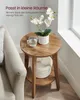 A round wooden side table with a plant and a glass of water on top, placed next to a sofa in a room with a window. Produktbild-Vorschau