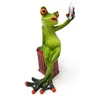 A decorative figurine of a frog taking a selfie with a cell phone, leaning on a suitcase. Produktbild-Vorschau