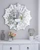 A decorative sunburst mirror hangs on a light gray wall above a white dresser. On the dresser are a few decorative items, including a bouquet of pink roses, a candle, and two glass bottles. A modern geometric lamp is also on the dresser. Náhľad obrázku produktu