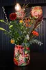A colorful vase filled with a bouquet of flowers sits on a dark surface, with a light bulb hanging above. Produktbild-Vorschau