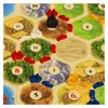 A close-up view of a board game, likely Settlers of Catan, showing hexagonal tiles with numbers and resource illustrations, along with game pieces. Náhled obrázku produktu