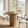 A round wooden dining table with chairs set in a bright room with large windows overlooking a garden. Produktbild-Vorschau