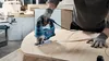 A person is using a blue Bosch jigsaw to cut a curved shape into a wooden surface in a workshop. Anteprima dell’immagine prodotto