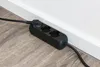 A black power strip with three outlets is plugged into a wall socket near the floor, resting on a wooden floor. Produktbild-Vorschau