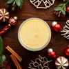 A jar of yellow liquid, likely Eierlikör, is surrounded by Christmas decorations like ornaments, pine sprigs, and cinnamon sticks on a wooden surface. Produktbild-Vorschau