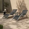 Two dark gray lounge chairs are set up on a gravel patio next to a small side table with a glass of water and a book. A potted olive tree is visible in the background, along with a wall casting dappled shadows. Voir les images du produit